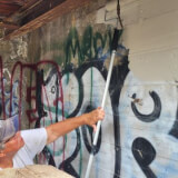 Woman using a roller to paint over graffiti on a concrete wall.